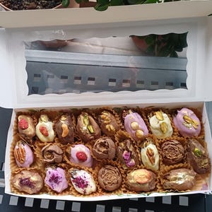 Puede incluir: Una caja blanca abierta con una ventana transparente, llena de dátiles variados cubiertos de chocolate. Los dátiles están decorados con nueces, frutas secas y pan de oro comestible. La caja está sobre una superficie oscura.