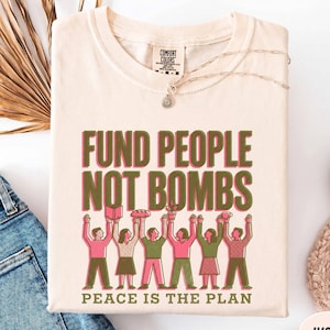K&ouml;nnte beinhalten: Elfenbeinfarbenes T-Shirt mit dem Text "FUND PEOPLE NOT BOMBS" in Gr&uuml;n und Rosa. Darunter eine Illustration von Personen, die Gegenst&auml;nde halten. Der Satz "PEACE IS THE PLAN" ist darunter gedruckt. Das Shirt wird mit blauen Jeans und einem wei&szlig;en Schuh pr&auml;sentiert.
