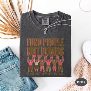 K&ouml;nnte beinhalten: Dunkelgraues T-Shirt mit dem Text "FUND PEOPLE NOT BOMBS" in Rosa und Gr&uuml;n. Das Shirt zeigt eine Grafik von Menschen, die Gegenst&auml;nde hochhalten. Der Satz "PEACE IS THE PLAN" ist unten aufgedruckt. Das Shirt ist auf einer wei&szlig;en Oberfl&auml;che gefaltet.