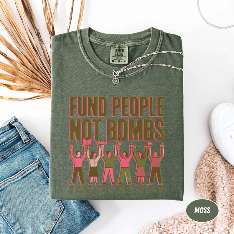 K&ouml;nnte beinhalten: Ein moosgr&uuml;nes T-Shirt mit dem Text "FUND PEOPLE NOT BOMBS" in Rosa und Braun. Die Grafik zeigt Menschen, die Gegenst&auml;nde hochhalten. Eine silberne Halskette und wei&szlig;e Turnschuhe sind ebenfalls zu sehen.