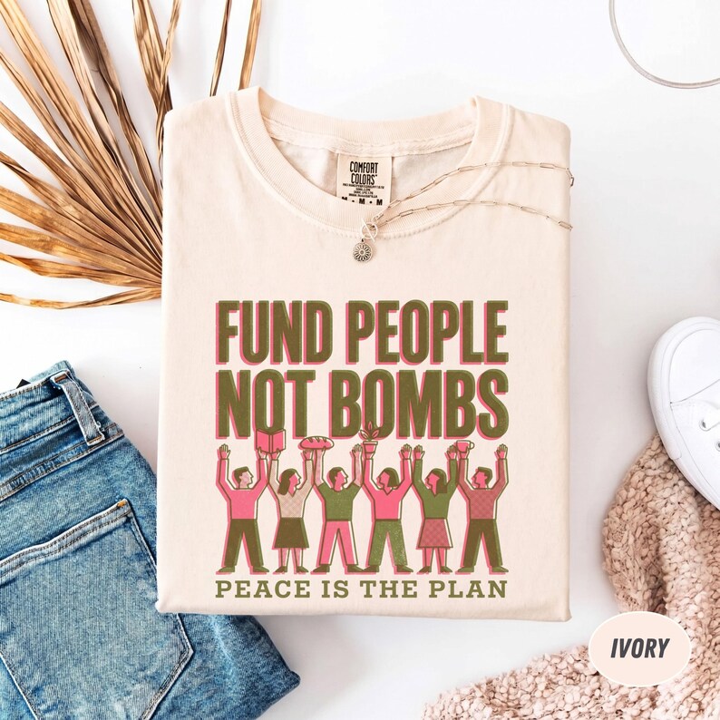 K&ouml;nnte beinhalten: Elfenbeinfarbenes T-Shirt mit dem Text "FUND PEOPLE NOT BOMBS" in Gr&uuml;n und Rosa. Darunter eine Illustration von Personen, die Gegenst&auml;nde halten. Der Satz "PEACE IS THE PLAN" ist darunter gedruckt. Das Shirt wird mit blauen Jeans und einem wei&szlig;en Schuh pr&auml;sentiert.
