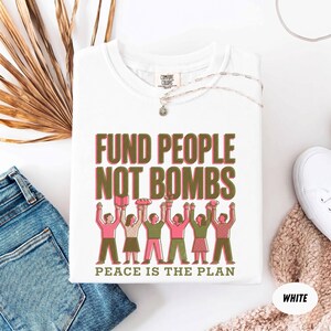 K&ouml;nnte beinhalten: Wei&szlig;es T-Shirt mit dem Text "FUND PEOPLE NOT BOMBS" in Rosa und Gr&uuml;n, mit einer Illustration von Personen, die Gegenst&auml;nde halten. Der Satz "PEACE IS THE PLAN" steht darunter. Das Shirt liegt neben einer blauen Jeans und wei&szlig;en Turnschuhen.