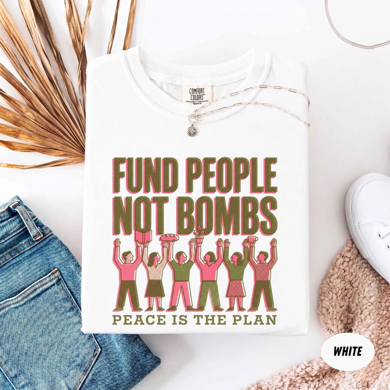 K&ouml;nnte beinhalten: Wei&szlig;es T-Shirt mit dem Text "FUND PEOPLE NOT BOMBS" in Rosa und Gr&uuml;n, mit einer Illustration von Personen, die Gegenst&auml;nde halten. Der Satz "PEACE IS THE PLAN" steht darunter. Das Shirt liegt neben einer blauen Jeans und wei&szlig;en Turnschuhen.