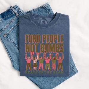 K&ouml;nnte beinhalten: Marineblaues T-Shirt mit dem Text "FUND PEOPLE NOT BOMBS" in Gr&uuml;n und Pink. Darunter eine Illustration von Menschen mit erhobenen Armen. Am unteren Rand steht "PEACE IS THE PLAN".