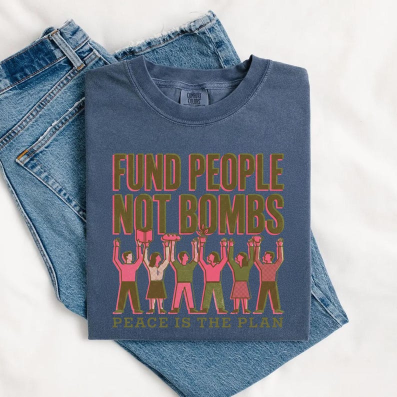 K&ouml;nnte beinhalten: Marineblaues T-Shirt mit dem Text "FUND PEOPLE NOT BOMBS" in Gr&uuml;n und Pink. Darunter eine Illustration von Menschen mit erhobenen Armen. Am unteren Rand steht "PEACE IS THE PLAN".