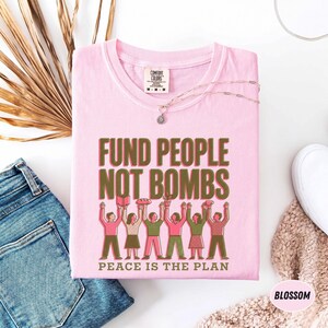 K&ouml;nnte beinhalten: Hellrosa T-Shirt mit dem Text "FUND PEOPLE NOT BOMBS" in Gr&uuml;n und Braun. Darunter steht "PEACE IS THE PLAN". Das Shirt zeigt eine Grafik von Menschen, die Gegenst&auml;nde halten. Eine silberne Halskette und eine Jeans sind ebenfalls zu sehen.