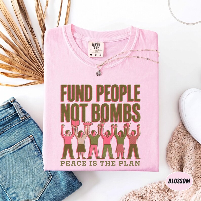 K&ouml;nnte beinhalten: Hellrosa T-Shirt mit dem Text "FUND PEOPLE NOT BOMBS" in Gr&uuml;n und Braun. Darunter steht "PEACE IS THE PLAN". Das Shirt zeigt eine Grafik von Menschen, die Gegenst&auml;nde halten. Eine silberne Halskette und eine Jeans sind ebenfalls zu sehen.