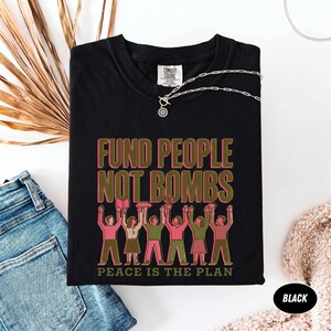 K&ouml;nnte beinhalten: Schwarzes T-Shirt mit dem Text "FUND PEOPLE NOT BOMBS" in Rosa und Gr&uuml;n. Darunter eine Illustration von Menschen mit erhobenen Armen. Der Satz "PEACE IS THE PLAN" ist darunter gedruckt. Eine silberne Halskette liegt auf dem T-Shirt.