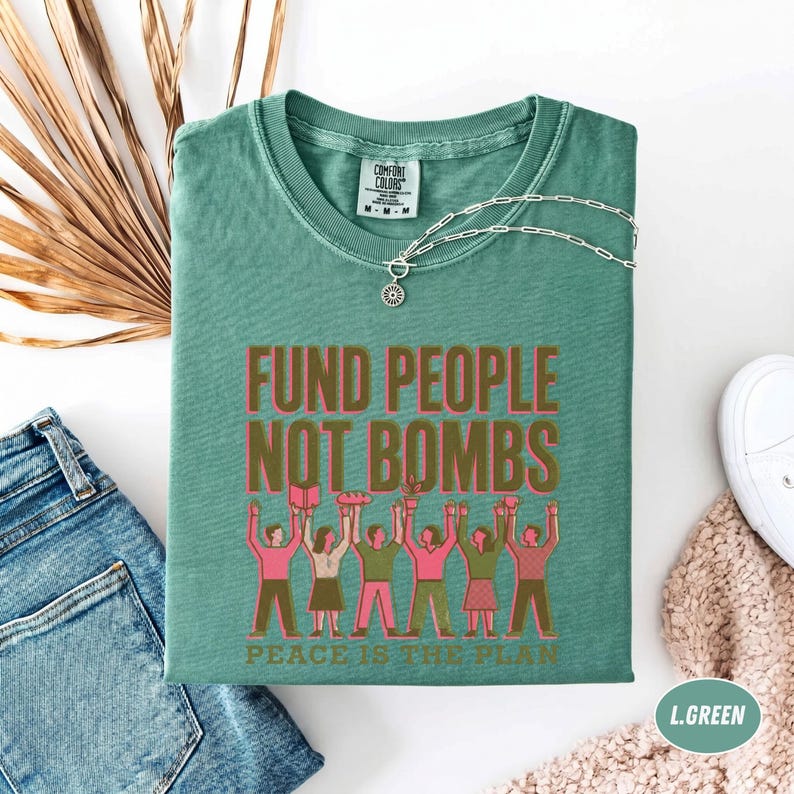K&ouml;nnte beinhalten: Ein gr&uuml;nes Comfort Colors T-Shirt mit dem Text "FUND PEOPLE NOT BOMBS" in Rosa und Braun. Das Shirt zeigt eine Grafik von Menschen, die Gegenst&auml;nde hochhalten. Eine silberne Halskette und eine blaue Jeans sind ebenfalls sichtbar.