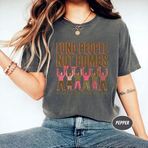 K&ouml;nnte beinhalten: Dunkelgraues T-Shirt mit dem Text "FUND PEOPLE NOT BOMBS" in Rosa und Gr&uuml;n. Darunter eine Illustration von Menschen, die die Arme heben. Der Satz "PEACE IS THE PLAN" ist unter der Illustration aufgedruckt.