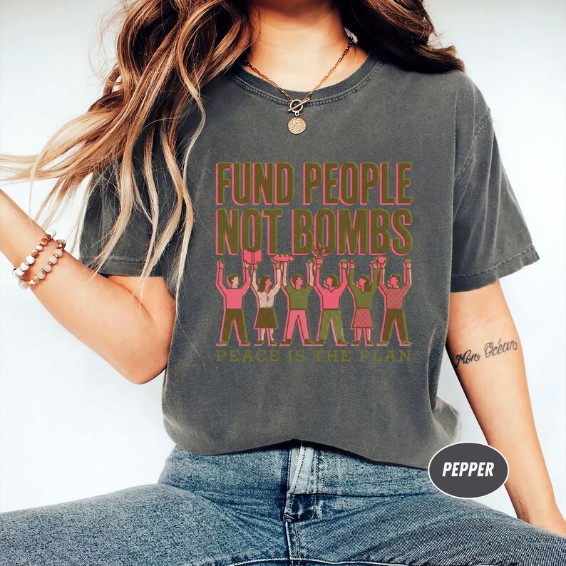 K&ouml;nnte beinhalten: Dunkelgraues T-Shirt mit dem Text "FUND PEOPLE NOT BOMBS" in Rosa und Gr&uuml;n. Darunter eine Illustration von Menschen, die die Arme heben. Der Satz "PEACE IS THE PLAN" ist unter der Illustration aufgedruckt.