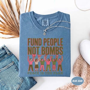 K&ouml;nnte beinhalten: Ein blaues Comfort Colors T-Shirt mit der Aufschrift "FUND PEOPLE NOT BOMBS" in Braun und Rosa. Die Grafik zeigt Personen, die Gegenst&auml;nde &uuml;ber ihren K&ouml;pfen halten. Der Text "PEACE IS THE PLAN" steht darunter. Eine silberne Halskette liegt auf dem Shirt.