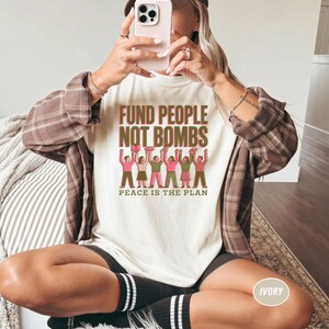 K&ouml;nnte beinhalten: Elfenbeinfarbenes T-Shirt mit dem Text "FUND PEOPLE NOT BOMBS" in Gr&uuml;n und Rot. Darunter steht "PEACE IS THE PLAN". Die Grafik zeigt Figuren mit erhobenen Armen. Die Person im Bild tr&auml;gt ein braunes kariertes Hemd.