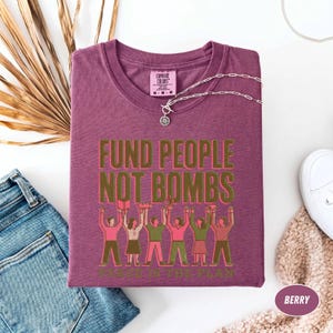 K&ouml;nnte beinhalten: Beerenfarbenes T-Shirt mit dem Text "FUND PEOPLE NOT BOMBS" in goldfarbenen Buchstaben. Das Shirt zeigt eine Grafik von Menschen, die Gegenst&auml;nde hochhalten. Eine silberne Halskette und blaue Jeans sind ebenfalls sichtbar.
