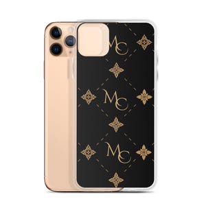 Op de afbeelding: Een goudkleurige smartphone met een doorzichtig beschermhoesje met een zwarte achtergrond, een herhalend gouden ruitpatroon, gouden bloemvormige iconen en de gouden letters MC gecentreerd in de ruitvormen op de achterkant.