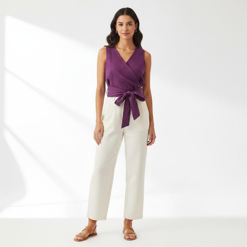 May include: A sleeveless purple wrap top featuring a V-neckline and a self-tie waist belt, paired with straight-leg off-white trousers. The ensemble is completed with simple brown flat sandals, creating a clean and coordinated outfit.