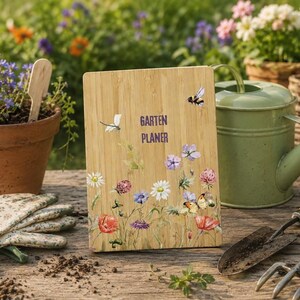 Könnte beinhalten: Ein Gartenplaner aus Holz mit dem Text "Garten Planer" und floralen und Schmetterlingsillustrationen. Der Planer ist hellbraun und zeigt eine Vielzahl bunter Blumen, darunter rote Mohnblumen, weiße Gänseblümchen und lila Blüten. Eine grüne Gießkanne und Gartengeräte befinden sich im Hintergrund.