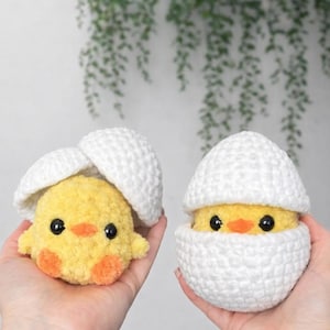 May include: Two handmade crochet chicks, one emerging from a white eggshell and the other inside. The chicks are yellow with orange feet and beaks, and black eyes. The eggshells are white. The background is a light color with green foliage.