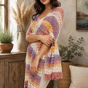 Peut inclure: Un châle en crochet multicolore aux tons rose, blanc, jaune et violet. Le châle a un col en V et un bord festonné. Le modèle porte le châle sur un haut blanc et un pantalon beige.