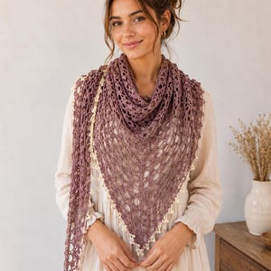 Könnte beinhalten: Ein gehäkelter Schal in Dreiecksform, in Mauve mit cremefarbenem Besatz. Der Schal ist über den Schultern drapiert und zeigt sein offenes Design und seine zarte Textur. Der cremefarbene Besatz bildet einen Kontrast.