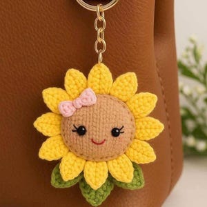 May include: A handmade crochet sunflower keychain with a smiling face and a pink bow. The sunflower has yellow petals, a brown center, and green leaves. The keychain is attached to a gold-colored metal ring.