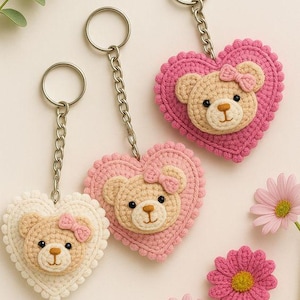 Op de afbeelding: Drie handgemaakte gehaakte sleutelhangers. Elke sleutelhanger heeft een hartvormig ontwerp met een teddybeergezicht en een kleine strik. De kleuren zijn roze, crème en beige, met zilverkleurige ringen en kettingen. Roze bloemen op de achtergrond.