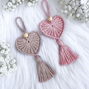 Op de afbeelding: Twee handgemaakte gehaakte hartornamenten. Eén is lichtroze, de andere is taupe. Elk hart heeft een houten kraal aan de bovenkant en een kwast aan de onderkant. De ornamenten worden gepresenteerd op een wit, pluizig oppervlak.