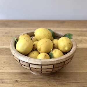 Può includere: Una ciotola di legno piena di limoni gialli freschi e alcune foglie verdi. La ciotola ha un design intrecciato unico e poggia su una superficie di legno. I limoni sono maturi e pronti per l'uso.