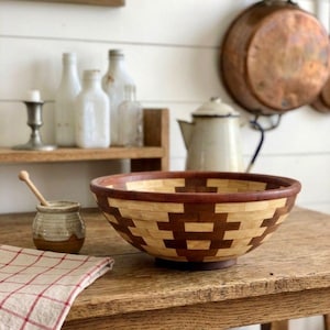 Può includere: Una grande ciotola decorativa in legno con un motivo geometrico nei toni del marrone e beige è appoggiata su un tavolo di legno rustico. La ciotola è circondata da altri oggetti da cucina, tra cui un piccolo barattolo con un cucchiaio di legno e un panno a scacchi rossi e bianchi.