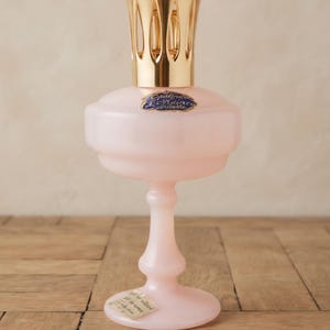 Pode incluir: Uma lâmpada de fragrância de vidro rosa pálido com uma base de pedestal e um corpo largo e arredondado. Possui um topo de metal dourado em forma de coroa e um rótulo oval azul na frente com o texto Opaline Lorraine. Um adesivo está na base.