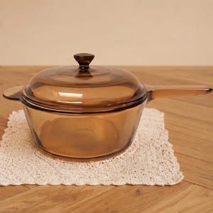 Pode incluir: Uma panela de vidro translúcido na cor âmbar com tampa a condizer e cabo comprido. O utensílio de cozinha apresenta um design liso e arredondado e está colocado sobre um tapete de croché quadrado em branco sujo com rebordo festonado.
