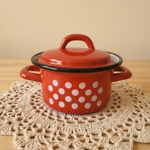 Pode incluir: Uma pequena panela redonda de esmalte vermelho com tampa a condizer e duas pegas laterais. O exterior da panela apresenta um padrão repetitivo de bolinhas brancas. O rebordo da panela e a tampa têm um acabamento com um rebordo preto.