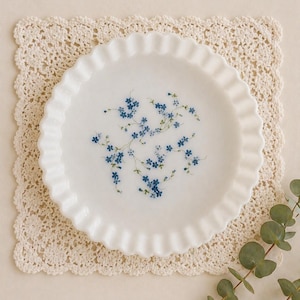 May include: A white, ruffled-edge plate with a delicate blue floral design, resting on a cream-colored crocheted doily. The plate is round and features a pattern of small blue flowers. A sprig of green leaves is in the lower right corner.