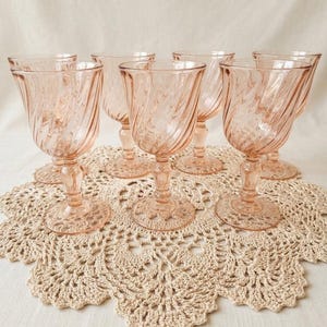 Peut inclure: Ensemble de six verres à vin roses avec un motif en spirale. Les verres sont disposés sur un napperon beige en crochet. La verrerie présente un design classique et élégant.