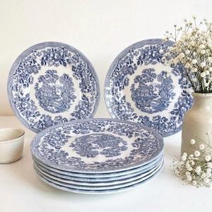 Peut inclure: Ensemble d'assiettes en céramique bleues et blanches avec un motif floral et paysager. Les assiettes sont empilées et disposées avec un petit bol crème et un vase de fleurs blanches. Les assiettes ont un bord festonné.
