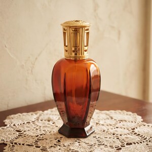 Op de afbeelding: Een geurlamp van doorschijnend amberkleurig glas met een gefacetteerde, zeshoekige voet en een afgerond lichaam. De bovenkant is voorzien van een gepolijste goudkleurige metalen branderkap met een decoratief uitgesneden ontwerp.