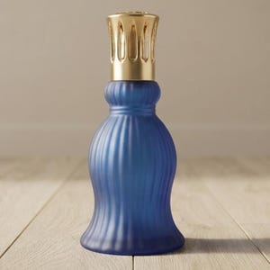 Może przedstawiać: Lampa zapachowa z niebieskiego matowego szkła o pionowej prążkowanej fakturze i szerokiej, rozszerzanej podstawie. Górna część lampy posiada złotą metalową nasadkę palnika z ozdobnymi wycięciami, kontrastującą z matowym niebieskim korpusem.