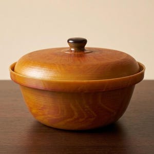 May include: A wooden bowl with a lid. The bowl and lid are a warm, reddish-brown colour with a wood grain pattern. The lid has a small, dark brown knob. The bowl sits on a dark brown surface.