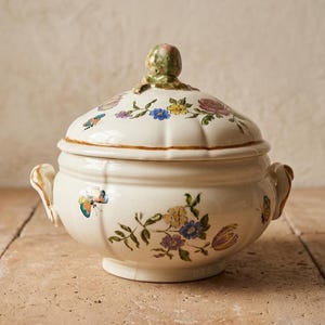 Peut inclure: Une soupière en céramique couleur crème avec un couvercle assorti doté d'un bouton sculpté. Le corps et le couvercle sont ornés de motifs floraux peints à la main dans des tons bleu, jaune et rose, parsemés de petites illustrations de papillons.