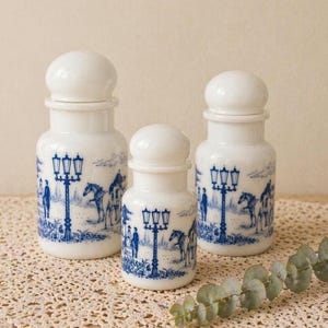 Può includere: Tre barattoli di ceramica bianca con coperchi arrotondati. Ogni barattolo presenta un disegno blu a trasferimento di una scena di strada con lampioni, figure e cavalli. I barattoli sono di diverse dimensioni, formando un set decorativo.