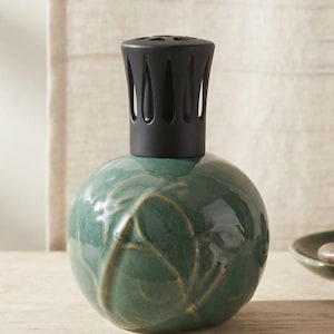 Peut inclure: Une lampe à parfum en céramique vert sauge de forme ronde avec une finition brillante et des motifs de feuilles en relief. Le récipient est surmonté d'un capuchon en métal noir mat avec des découpes verticales pour la diffusion.