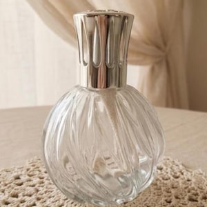 Peut inclure: Lampe à parfum en verre transparent avec un dessus en métal argenté. Le corps en verre rond présente un motif tourbillonnant texturé. La lampe repose sur une surface crème à tissage ouvert. La lampe est conçue pour diffuser des huiles parfumées.