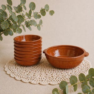 Può includere: Una collezione di ciotole color terracotta, tra cui una pila di sei e una ciotola più grande con manici. Sono appoggiate su un centrino all'uncinetto color crema. Foglie di eucalipto visibili sullo sfondo.