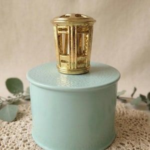 Op de afbeelding: Een cilindrische lichtblauwe keramische pot met een getextureerd deksel, bekroond met een decoratieve, goudkleurige metalen kooi-achtige knop. De houder heeft een gladde, effen gekleurde basis en een gedetailleerde, sierlijke gouden top.