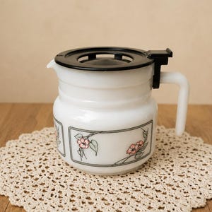 Può includere: Una caraffa da caffè in vetro opalino bianco con coperchio in plastica nera e manico bianco. Il corpo della caraffa è decorato con un bordo rettangolare contenente un motivo floreale di fiori rosa e foglie verdi su sfondo grigio chiaro.