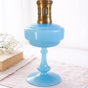 Könnte beinhalten: Eine hellblaue Glas-Öllampe mit einem Messingbrenner. Die Lampe hat einen abgerundeten Sockel, einen schlanken Stiel und einen bauchigen Behälter. Der Messingbrenner sitzt auf dem Behälter. Die Lampe steht auf einem neutralfarbenen Tuch.