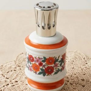 May include: A decorative ceramic oil lamp with a silver metal top. The white body features a floral pattern in red, orange, and pink. Orange bands accent the top and base. The lamp sits on a beige, crocheted doily.