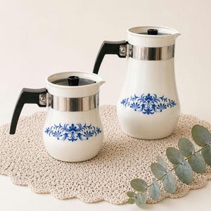 Peut inclure: Deux cafetières en céramique blanche avec des poignées et des couvercles noirs. Chaque pot a une bande argentée et un motif floral bleu. Les pots sont sur un dessous de plat beige crocheté.