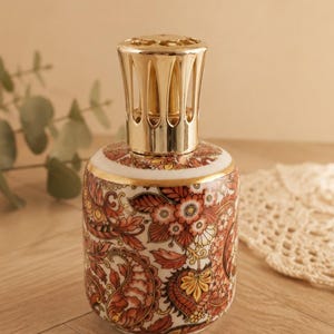 Op de afbeelding: Een cilindervormige keramische geurlamp met een gedetailleerd paisley- en bloemenpatroon in oranje, rode en gele tinten op een witte achtergrond. De lamp is afgewerkt met een glanzende goudkleurige metalen kroon met verticale uitsparingen.