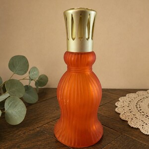 May include: A frosted orange glass fragrance lamp with a curved, ribbed body and a gold-toned metal vented cap. The vessel features a wide base that tapers into a narrow neck, topped with a decorative metallic crown designed for air diffusion.